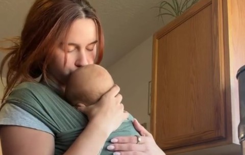 O tânără care sărută blând pe cap nou-născutul purtat într-un sistem de wrap textil verde, într-un cadru casnic.