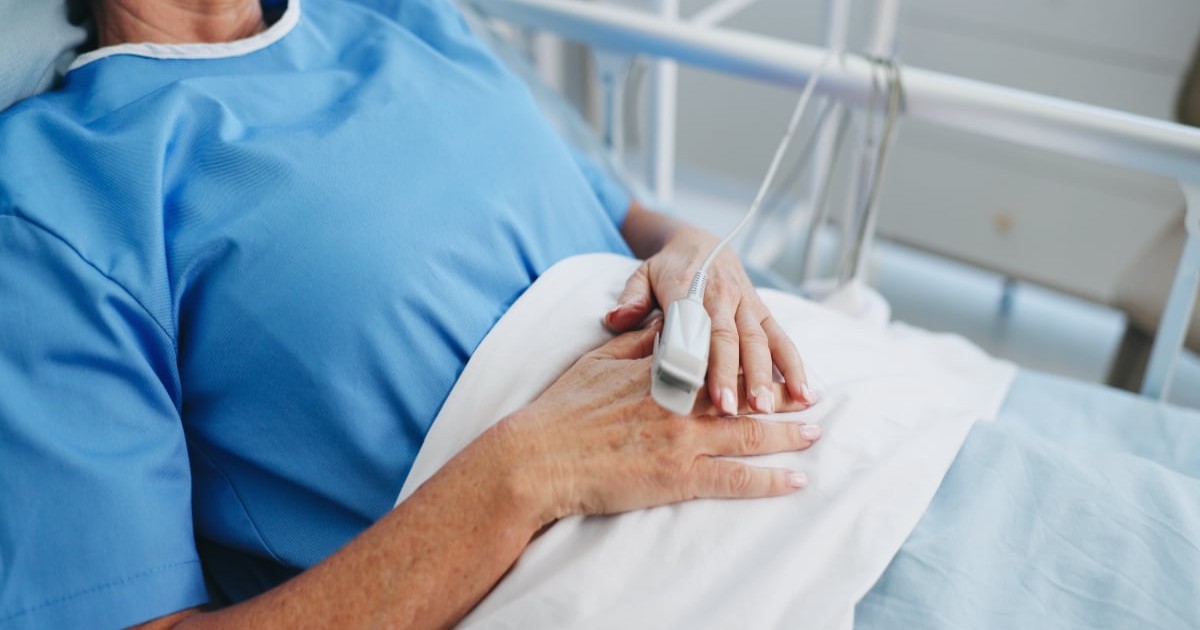 Imaginea arată o pacientă internată în spital, întinsă pe pat, cu un senzor medical montat pe deget pentru monitorizarea funcțiilor vitale.