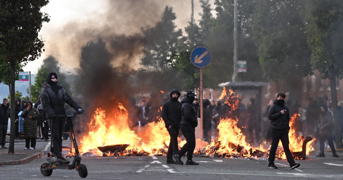 Mai mulți tineri strânși în jurul unei baricade de foc în mijlocul unei străzi, în timpul protestelor din Belfast