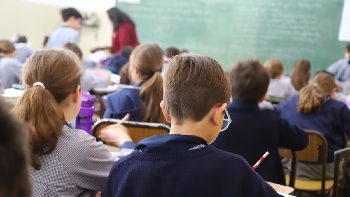 Elevi care stau în bănci și scriu. În fundal se observă o femeie cu pulover roșu