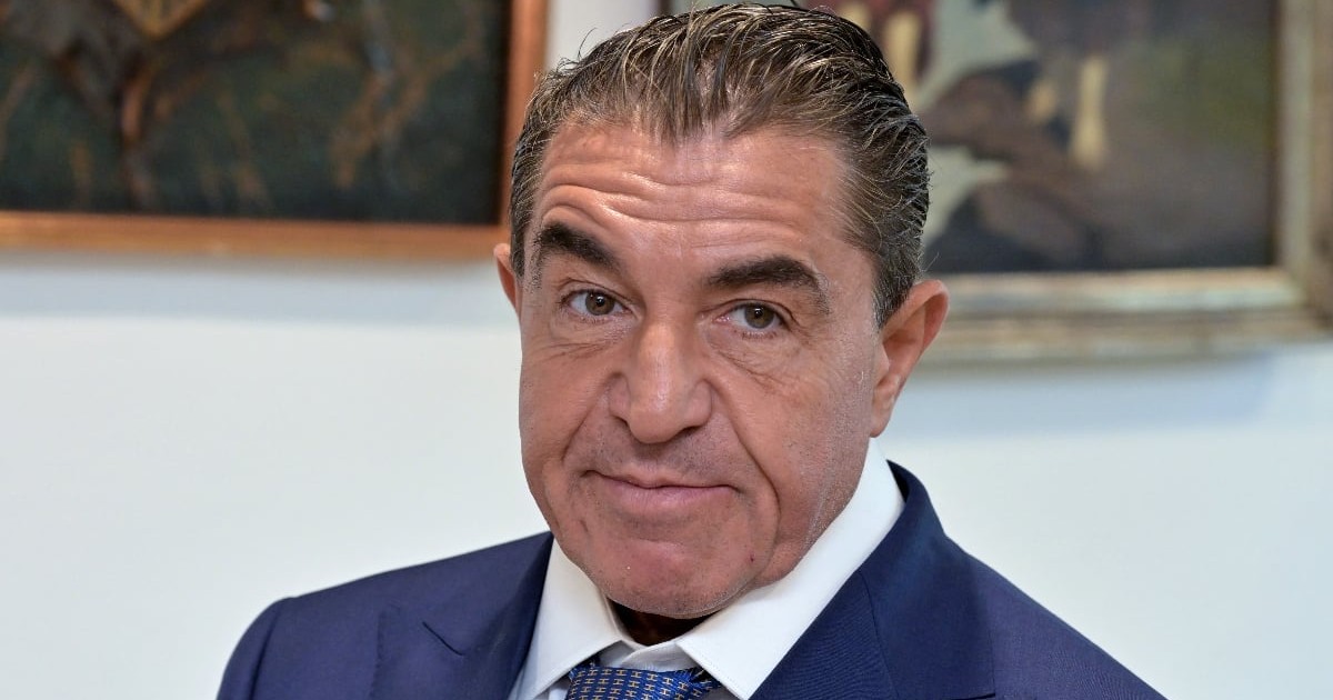 Un bărbat îmbrăcat elegant, în costum și cravată, privește direct către cameră. Fundalul sugerează un interior formal, cu tablouri expuse pe perete.