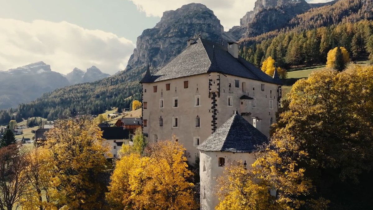 Fotografie cu un castel medieval din piatră, situat într-o pădure luxuriantă de toamnă, cu copaci aurii, înconjurat de munți și un cer parțial înnorat.