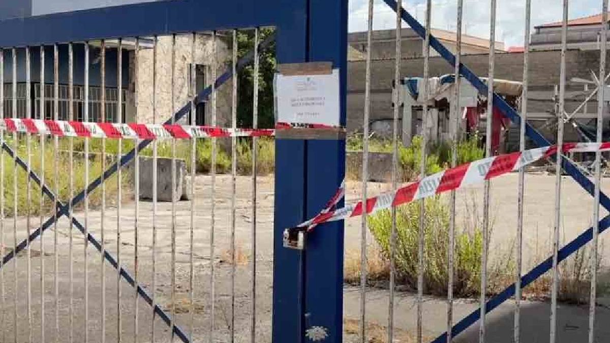 O poartă legată și securizată cu bandă a poliției, în spatele ei clădiri abandonate