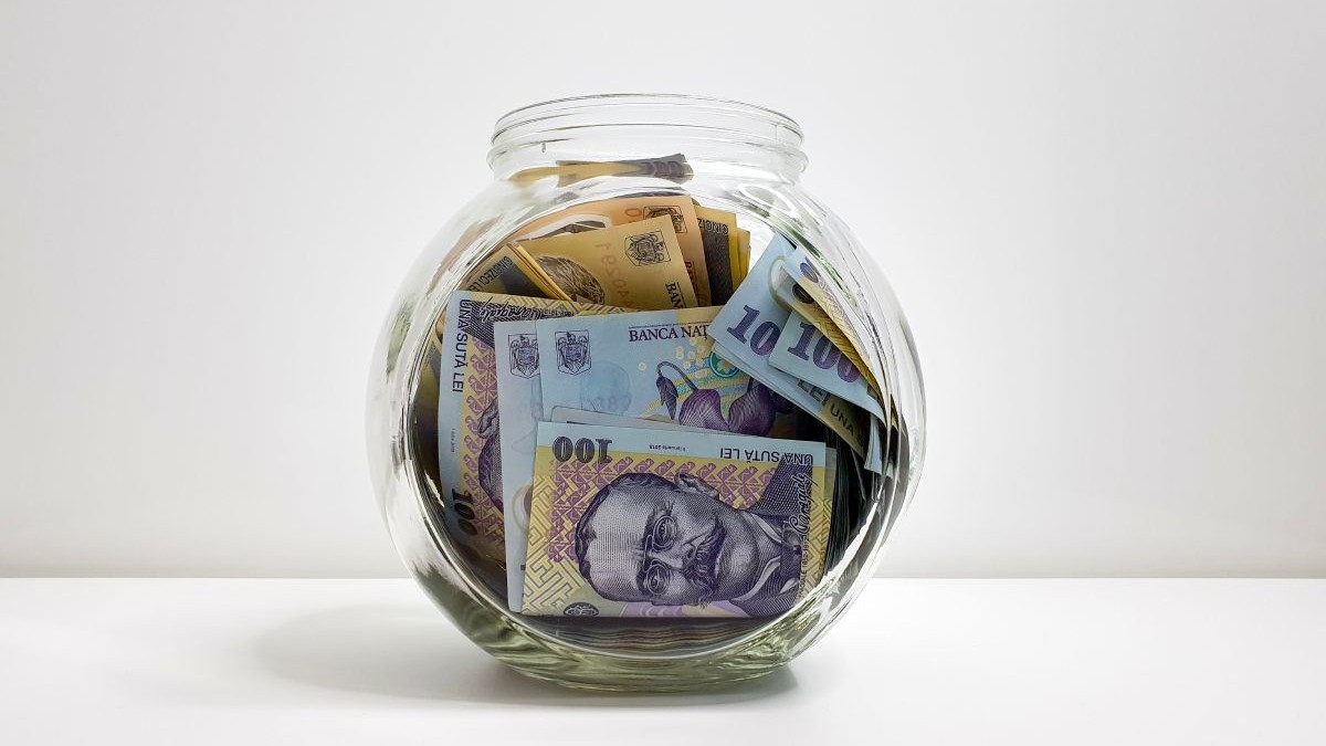 Borcan din sticlă transparentă în care se află bancnote de 100 de lei, pe o masă albă, pe fundal alb