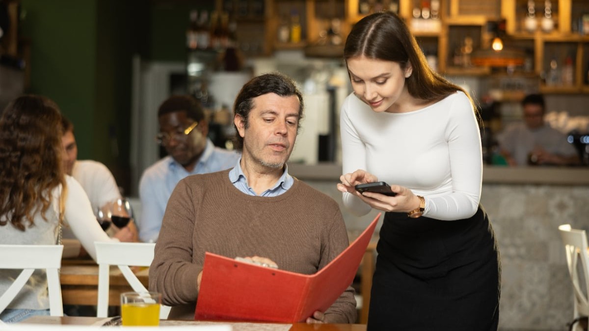 Un client care stă la masă într-un restuarant, cu meniul în mână, în timp ce ospătărița îi ia comanda