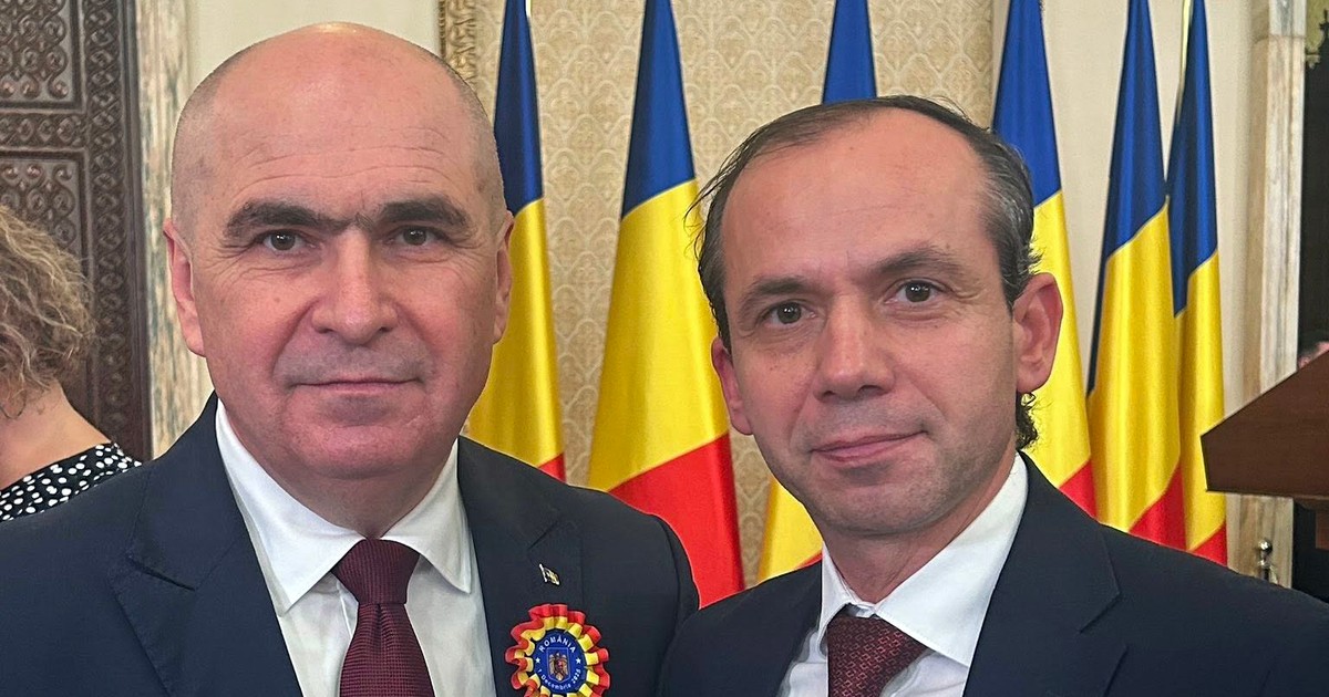Ilie Bolojan, premierul României, imbracat in costum negru, alături de primarul orasului Cahul, Republica Moldova, Imbracat in costum negru. In fundal se vad steaguri in rosu, galben si albastru