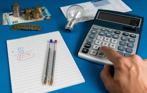 O factură de electricitate cu un bec, o mână calculează factura folosind un calculator și un caiet cu două pixuri și multe monede euro pe fundal albastru, iar sub ele este o hârtie cu un calcul și două pixuri, unul albastru și unul roșu. Foto: Shutterstock