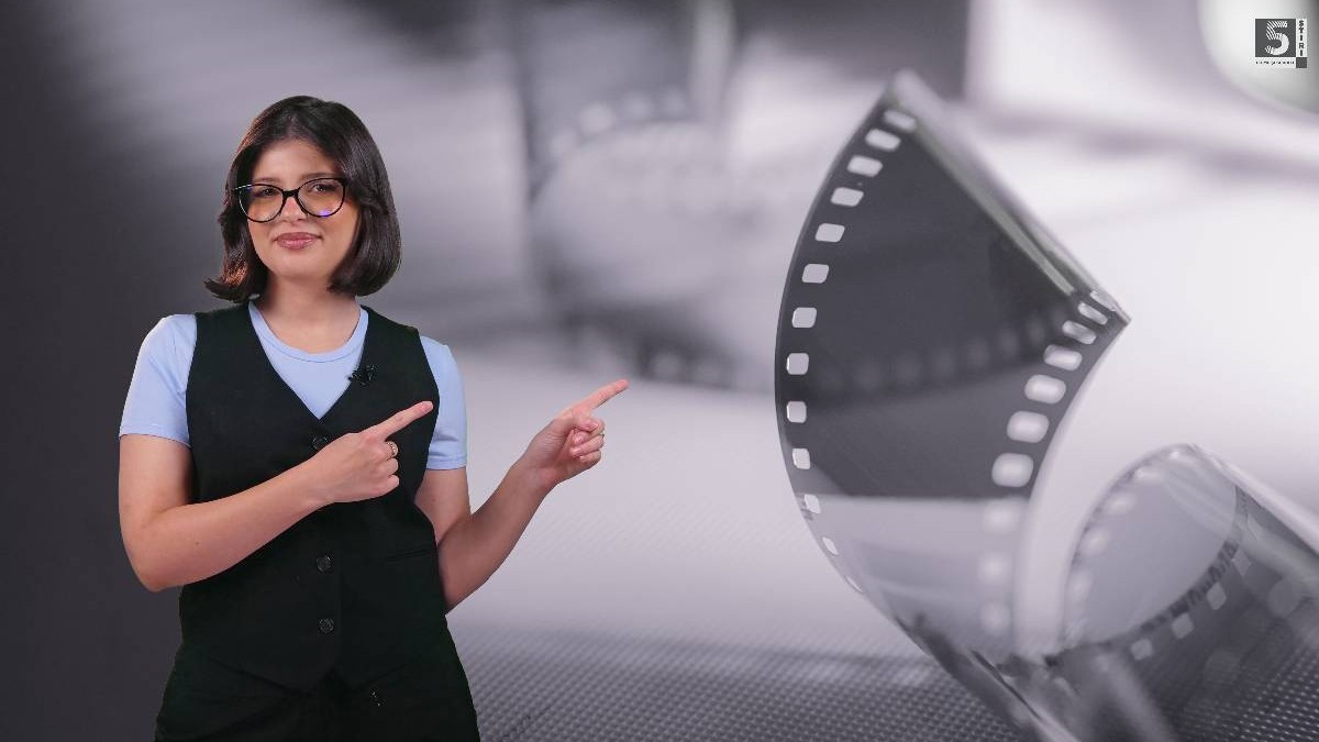 În imagine, Adelina Duinea, îmbrăcată într-un sarafan negru, cu o bluză albastră. Pe fundal e o poză cu o rolă de film