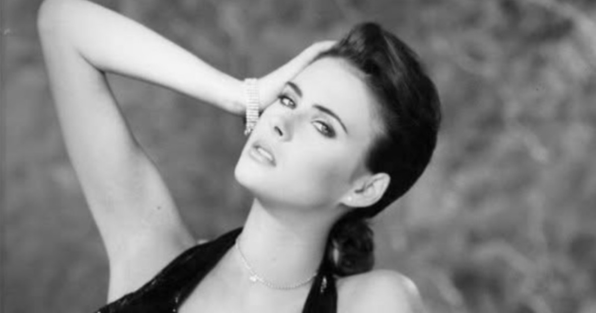 Fotografia alb-negru prezintă o femeie pozată într-un stil elegant, cu părul prins și o mână ridicată la ceafă. Ea poartă un top cu bretele subțiri și bijuterii delicate, iar imaginea are un aer clasic de ședință foto de modă.
