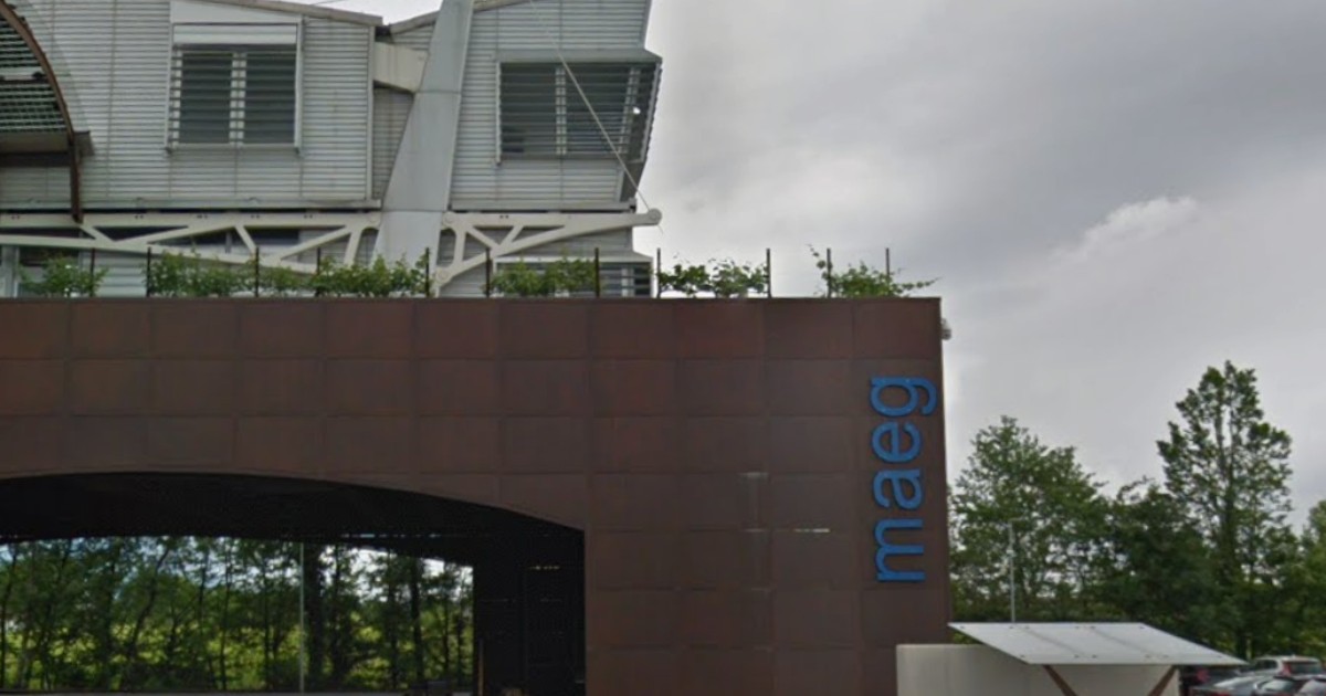 Imaginea prezintă sediul unei companii moderne, cu o fațadă metalică de culoare brun-ruginie și sigla „maeg” afișată vizibil pe clădire. Construcția are un design industrial contemporan, cu elemente arhitecturale din metal și sticlă, fiind amplasată într-o zonă înconjurată de vegetație și locuri de parcare.