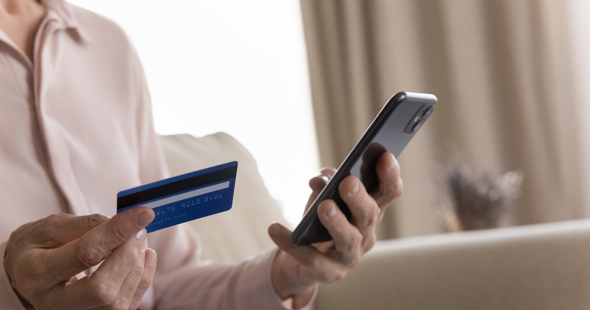Imaginea prezintă o persoană care ține în mână un card bancar și un telefon mobil, sugerând efectuarea unei operațiuni financiare online. Fotografia este ilustrativă și face trimitere la utilizarea aplicațiilor de mobile banking și a plăților digita