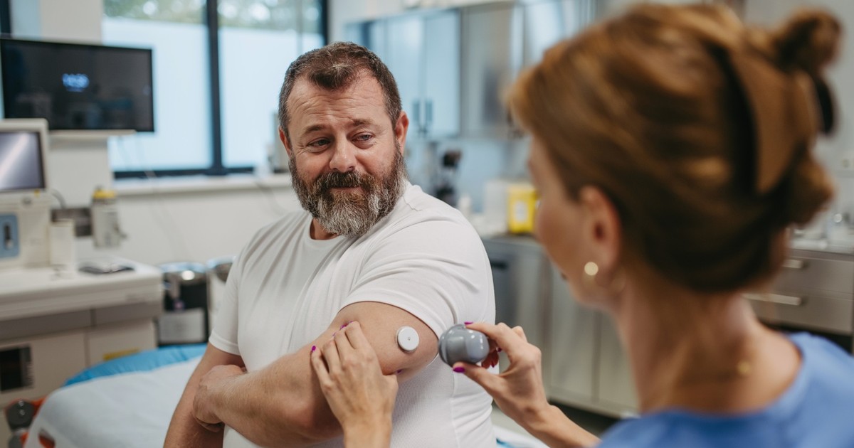 Un barbatcu barba, imbracat in tricou alb, cu senzor glicemie pe brat, sta pe un pat intr-un cabinet medical, iar o femeie cadru medical verifica senzorul punand mana pe brat