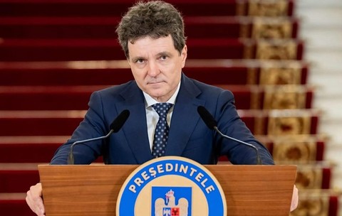 Nicușor Dan îmbrăcat în costum bleumarin și cravată cu model, privind spre cameră din spatele unui pupitru oficial.
