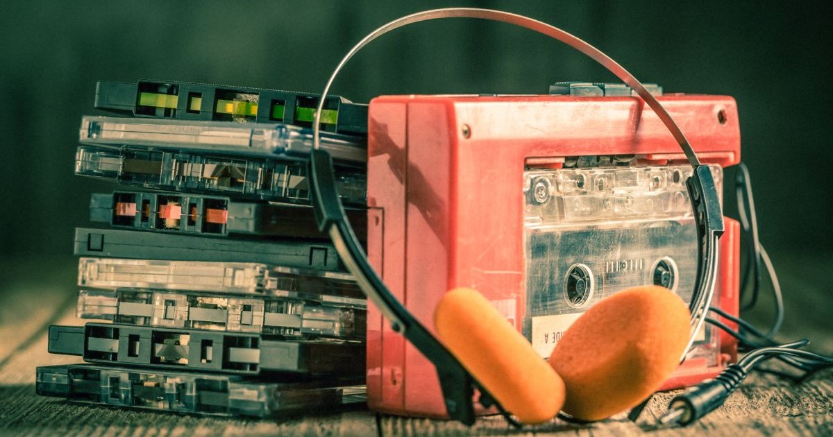 Casete audio cu muzică, așezate una peste alta, cu un walkman de culoare roșie lângă, pe o masă, și o pereche de căști cu burete de culoare portocalie