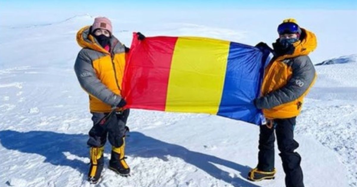două tinere îmbrăcate în echipament de escaladare montană, format din căciulă, geci portocalii, pantaloni negrii și cizme negre cu dungi portocalii, ținând în mâini drapelul României, pe vârful unui munte