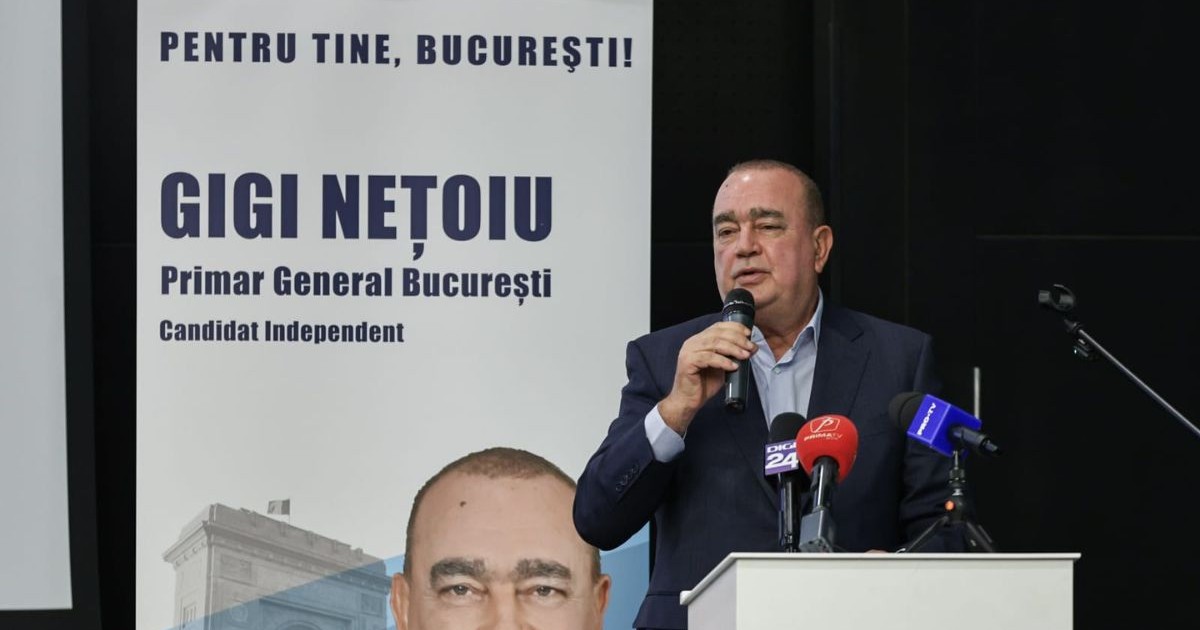 Gigi Nețoiu, vorbind la microfon la o conferință de presă, în fața unui banner cu numele său