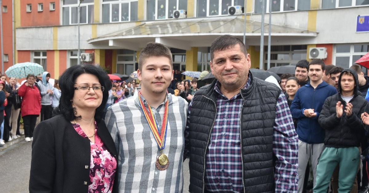 Un adolescent cu cămasa in carouri și medalii la gât, pozează alături de o femeie cu bluza roz si un bărbat care poarta o cămasa in carouri, in curtea unui liceu