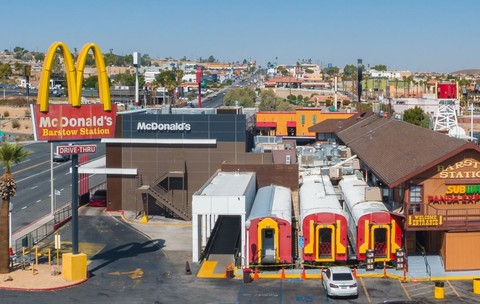 Vedere aeriană a unui centru comercial plin de viață din Barstow, cu restaurante emblematice precum McDonald's, pe fundalul munților, al cerului senin și al unui tren care trece.
