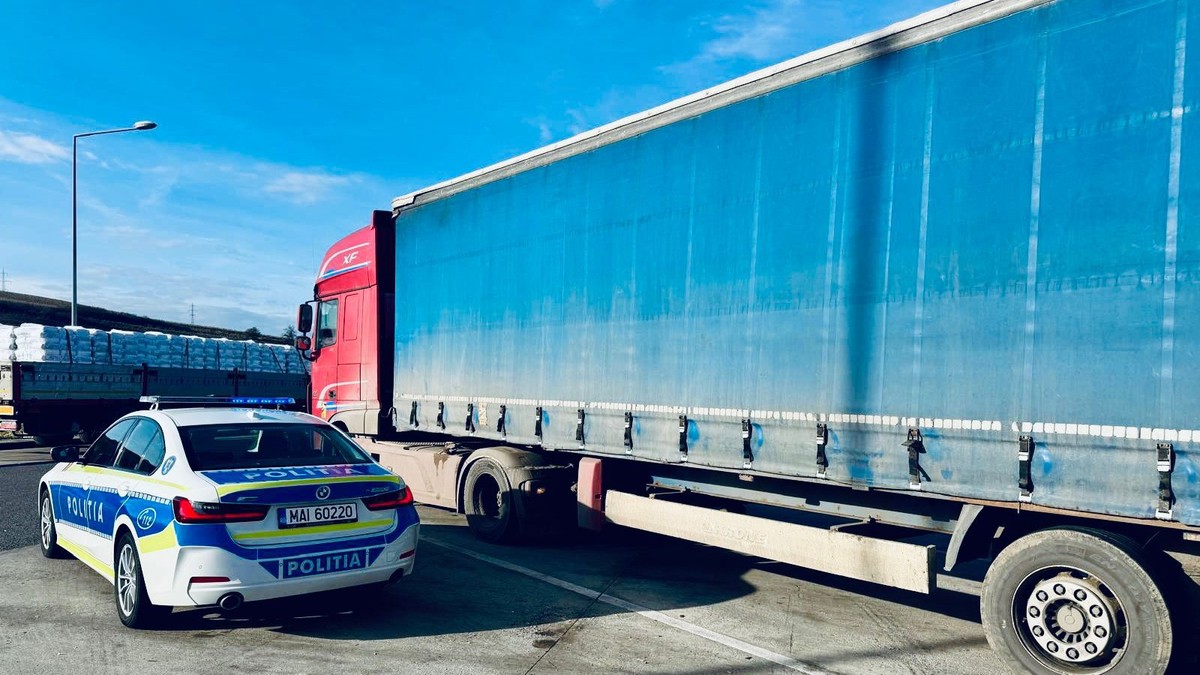Fotografia surprinde omașină de poliție parcată în stânga, lângă un camion mare de marfă albastru în dreapta.