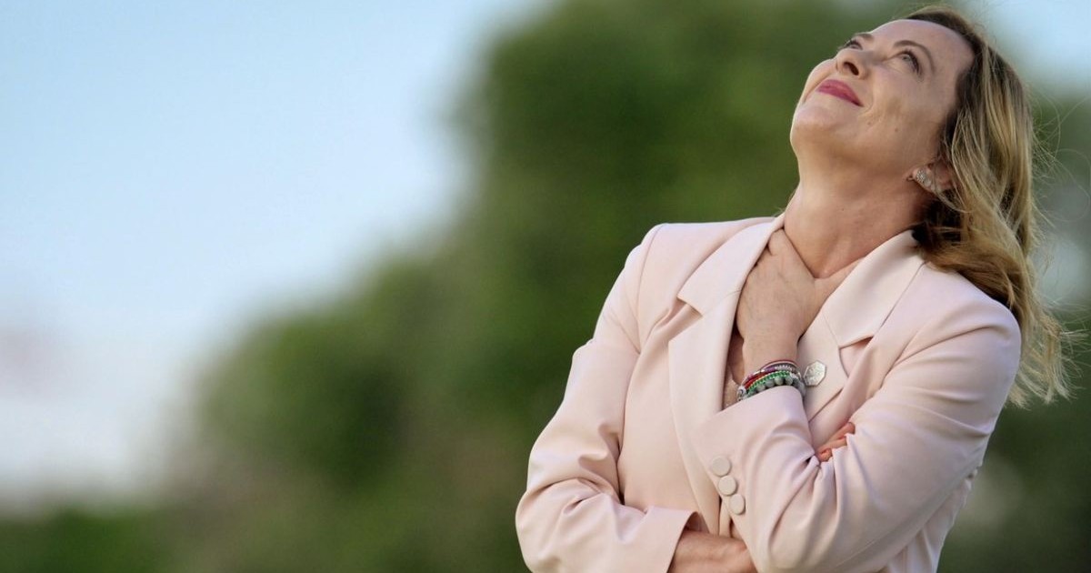 Premierul italian Georgia Meloni în mijlocul naturii, îmbrăcată într-un costum roz piersică, cu capul pe spate, părul fluturând si mâna stangă pusă la gat