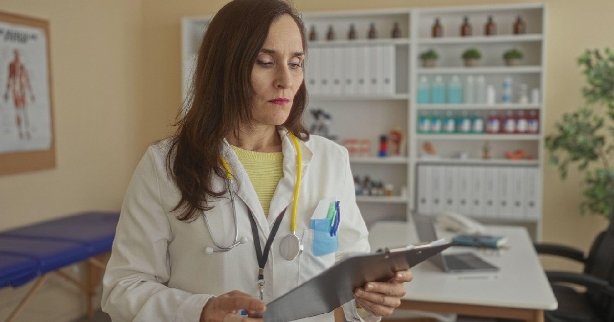Medic femeie în uniformă, echipată cu stetoscop și clipboard, analizând atent dosarele pacienților într-un cabinet medical, înconjurată de echipamente și materiale sanitare