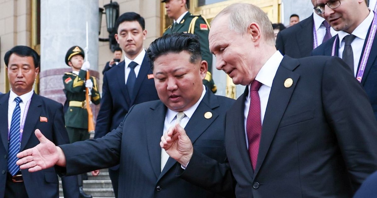 Vladimir Putin zambind, dreapta, alaturi de Kim Jong Un,, care are mana dreapta intinsa inainte, stanga, incongurati de mai multi barbati din echipa de securitate