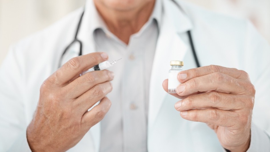 Un doctor îmbrăcat în halat alb, purtând un stetoscop în jurul gâtului, care ține o seringă cu ac și un flacon mic de sticlă