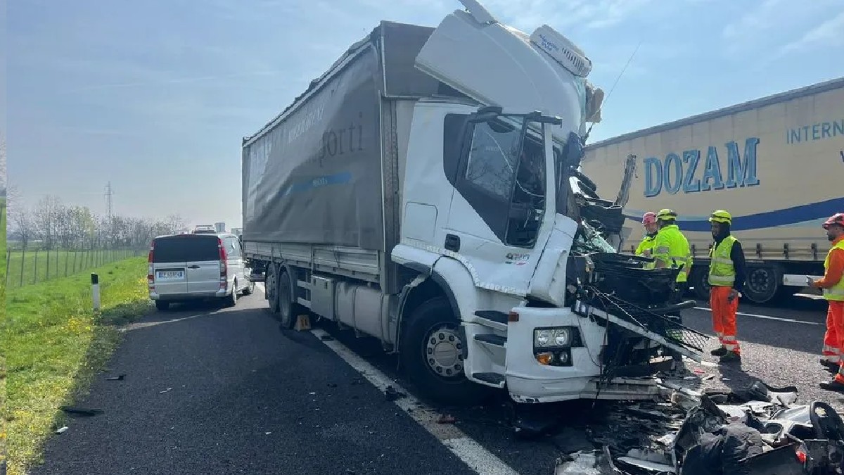 Imaginea surprinde urmările unui accident rutier grav pe o autostradă, în care un camion alb cu cabina puternic avariată este oprit pe marginea drumului, fiind înconjurat de echipaje de intervenție și resturi de caroserie împrăștiate pe carosabil.