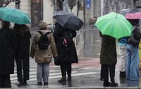 Mai mulți pietoni așteptând la o trecere de pietoni pe o vreme ploioasă și rece, adăpostindu-se sub umbrele colorate și purtând haine groase.