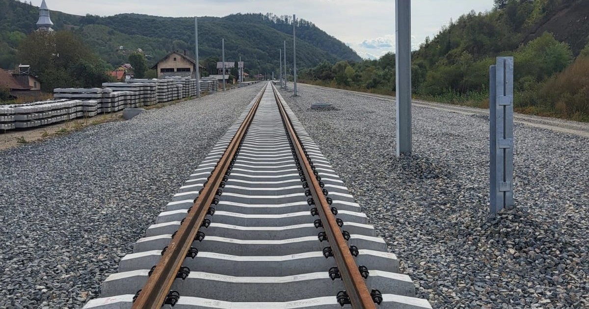 Imaginea surprinde un tronson de cale ferată aflat în curs de modernizare, cu șine noi montate pe traverse de beton, într-o zonă deluroasă. În jur se văd materiale de construcție și stâlpi instalați, semn că lucrările sunt încă în desfășurare.