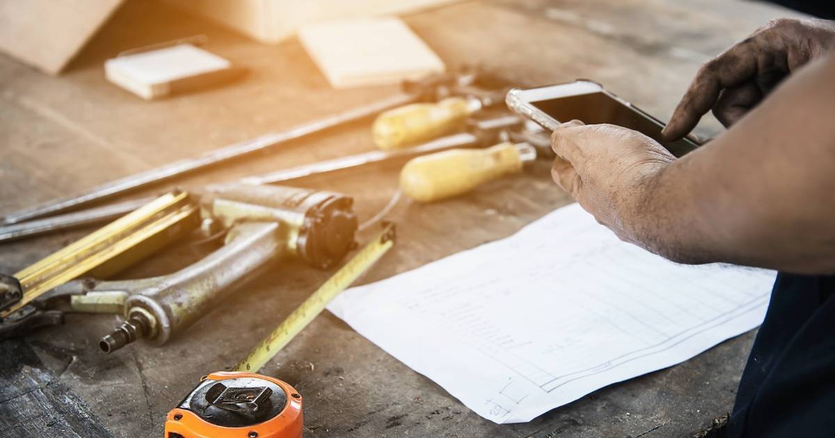 Dulgher care lucrează la pregătirea produselor de mobilier din lemn folosind unelte manuale și o stație de lucru pe masă, în timp ce folosește telefonul mobil