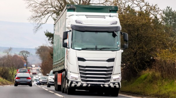 Un camion alb deplasându-se pe o șosea alături de alte autoturisme, într-o zonă de drum mărginită de vegetație.
