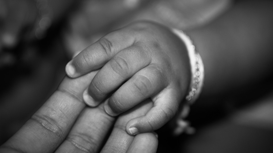 Imaginea alb-negru prezintă un prim-plan cu mâna mică a unui copil care își ține degetele peste palma unui adult.