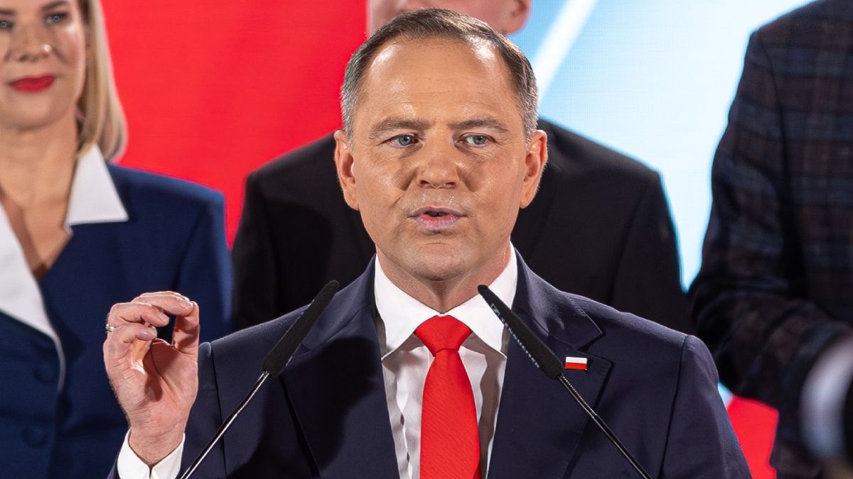 Karol Nawrocki, in sacou negru si cravata rosie, vorbeste la microfon cu mana dreaptă ridicată si oameni din staff în spate