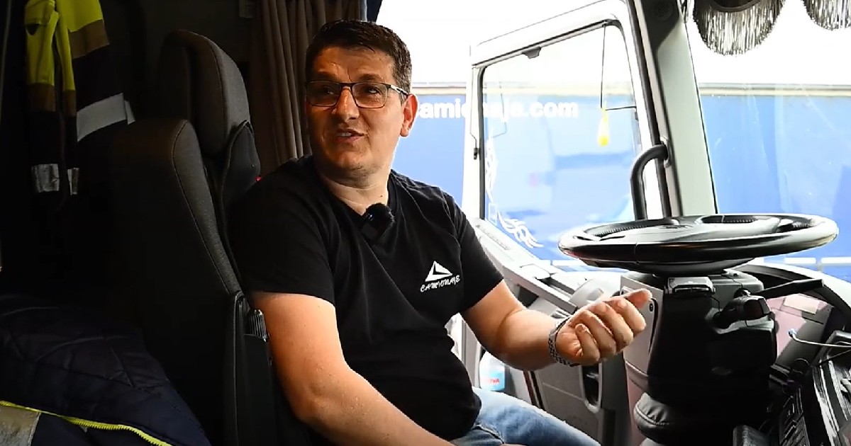 Imaginea prezintă un bărbat cu ochelari și tricou negru stând pe scaunul șoferului în cabina unui camion, surprins în timp ce gesticulează și vorbește.