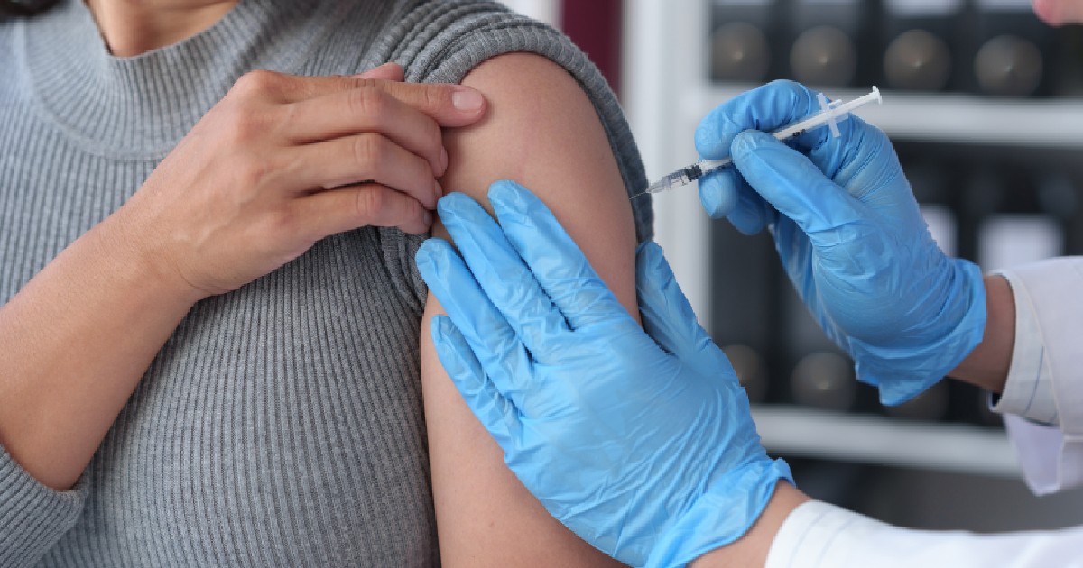 Imaginea surprinde de aproape momentul administrării unui vaccin sau a unei injecții într-un cadru medical. O persoană îmbrăcată în gri își ține brațul, în timp ce o mână a unui lucrător medical, protejată de mănuși albastre, injectează serul cu o seringă.