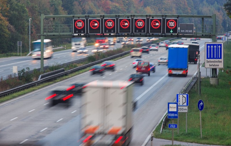Imaginea prezintă traficul de pe o autostradă din Germania (Autobahn), unde panouri electronice deasupra benzilor afișează o limită de viteză de 100 km/h și restricții de depășire pentru camioane.