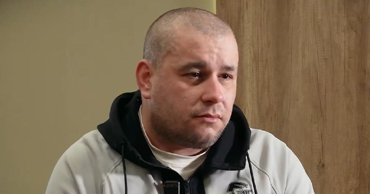 Un bărbat chel, cu o expresie serioasă, îmbrăcat într-o jachetă gri cu negru, cu lacrimi în ochi, stă în fața unui perete bej și a unei uși din lemn.