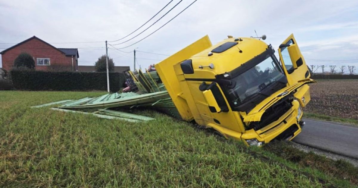 Imaginea arată un camion galben răsturnat pe o parte într-un câmp. În fundal se vede o casă din cărămidă roșie cu acoperișul închis la culoare.