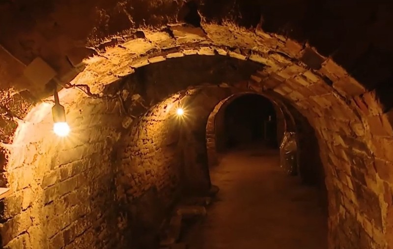 Interiorul unui tunel sau al unui beci subteran vechi, zidit din cărămidă roșie cu tavane boltite, iluminat de becuri.