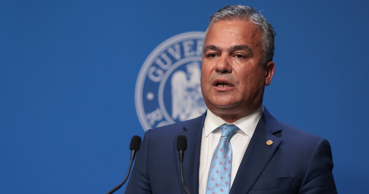 Imaginea prezintă un bărbat îmbrăcat într-un costum închis la culoare și cămașă albă, cu o cravată bleu deschis, surprins într-un cadru oficial. Expresia serioasă și postura sobră sugerează participarea la un eveniment instituțional sau politic.