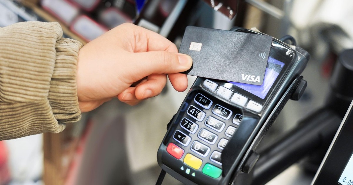 Mâna unui client ține un card de debit Visa negru lângă un terminal de plată pentru o tranzacție de plată fără contact.