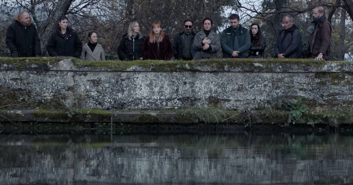 Un grup de doisprezece oameni care stau pe marginea unui zid de piatră și privesc spre un lac.