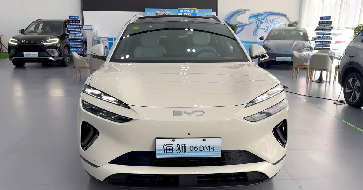 Prim-plan cu partea frontală a unei mașini BYD Sealion 06 DM-i, albă, într-un show-room auto, cu alte autoturisme în fundal