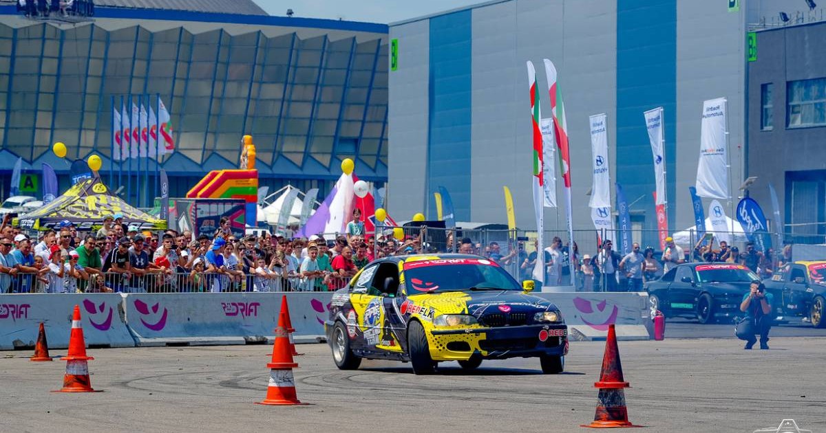 demonstrație de drift, desfășurată în fața unui public numeros, în cadrul unui eveniment auto organizat la Romexpo. În prim-plan, un BMW modificat, decorat cu stickere viu colorate, derapează controlat printre jaloane portocalii, lăsând urme de cauciuc pe asfalt. Pe fundal, zeci de spectatori privesc momentul