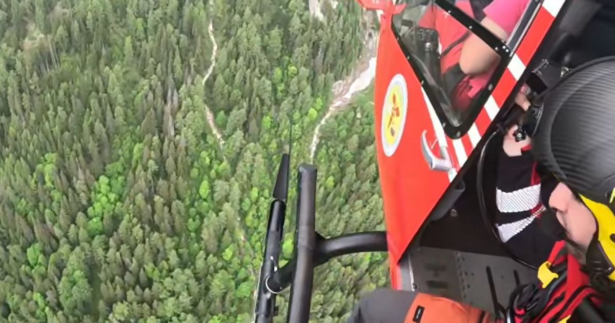 Imagini din elicopter cu salvamontist imbracat in rosu care se pregateste de coborare pe troliu pentru o operațiune de salvare deasupra unei paduri verzi