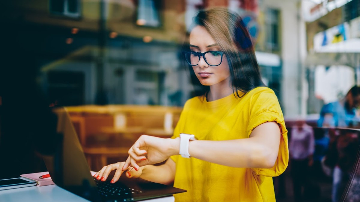 Femeie tanara, lucreaza la laptop intr-o cafenea, isi verifica smartwatchul de la mana