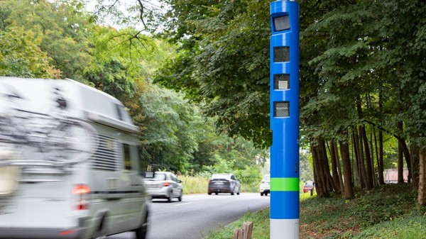 Un stâlp cilindric albastru înalt, prevăzut cu camere de supraveghere și senzori, amplasat pe marginea unei șosele mărginite de copaci în timp ce trec vehicule.