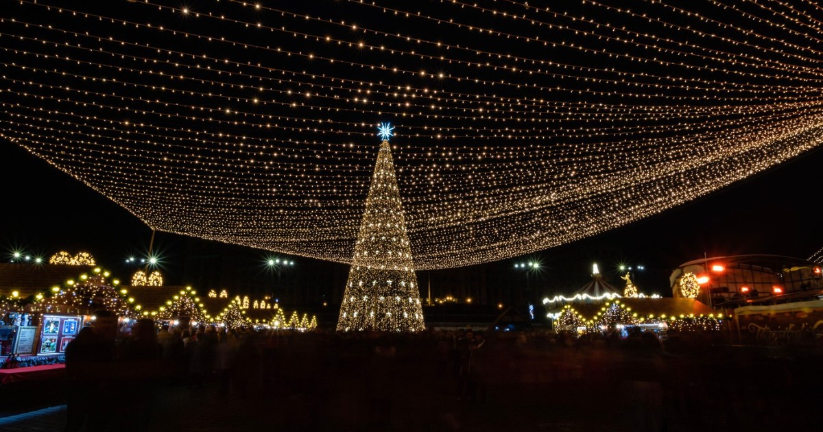 Imaginea surprinde un târg de Crăciun luminat spectaculos, cu un brad impozant decorat și o boltă de lumini care acoperă întreaga piață. Atmosfera festivă este completată de mulțimea de vizitatori și de standurile strălucitoare, care creează un cadru de sărbătoare și bucurie.