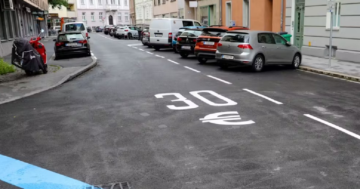 O stradă asfaltată, cu mașini parcate pe partea dreaptă, iar pe carosabil este pictat mare cu alb „30” alături de simbolul euro, lângă marcaje de parcare trasate cu linii albe.